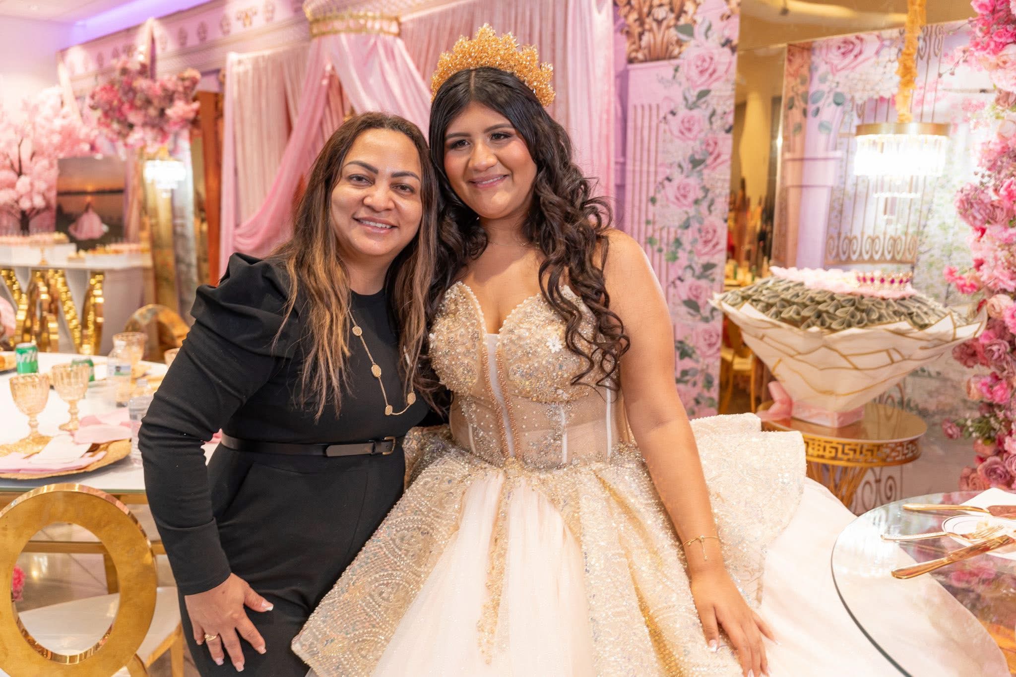 Two women smiling at a celebration with pink and gold decorations, one in a black dress and the other in a gold gown with a crown.