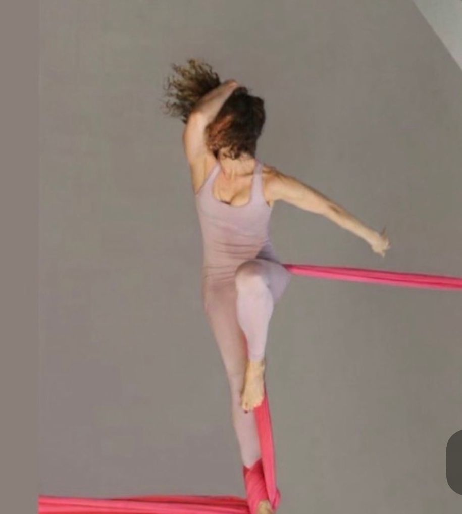 A person performing aerial silk acrobatics, hanging horizontally with one arm extended upward, on pink fabric against a gray background.