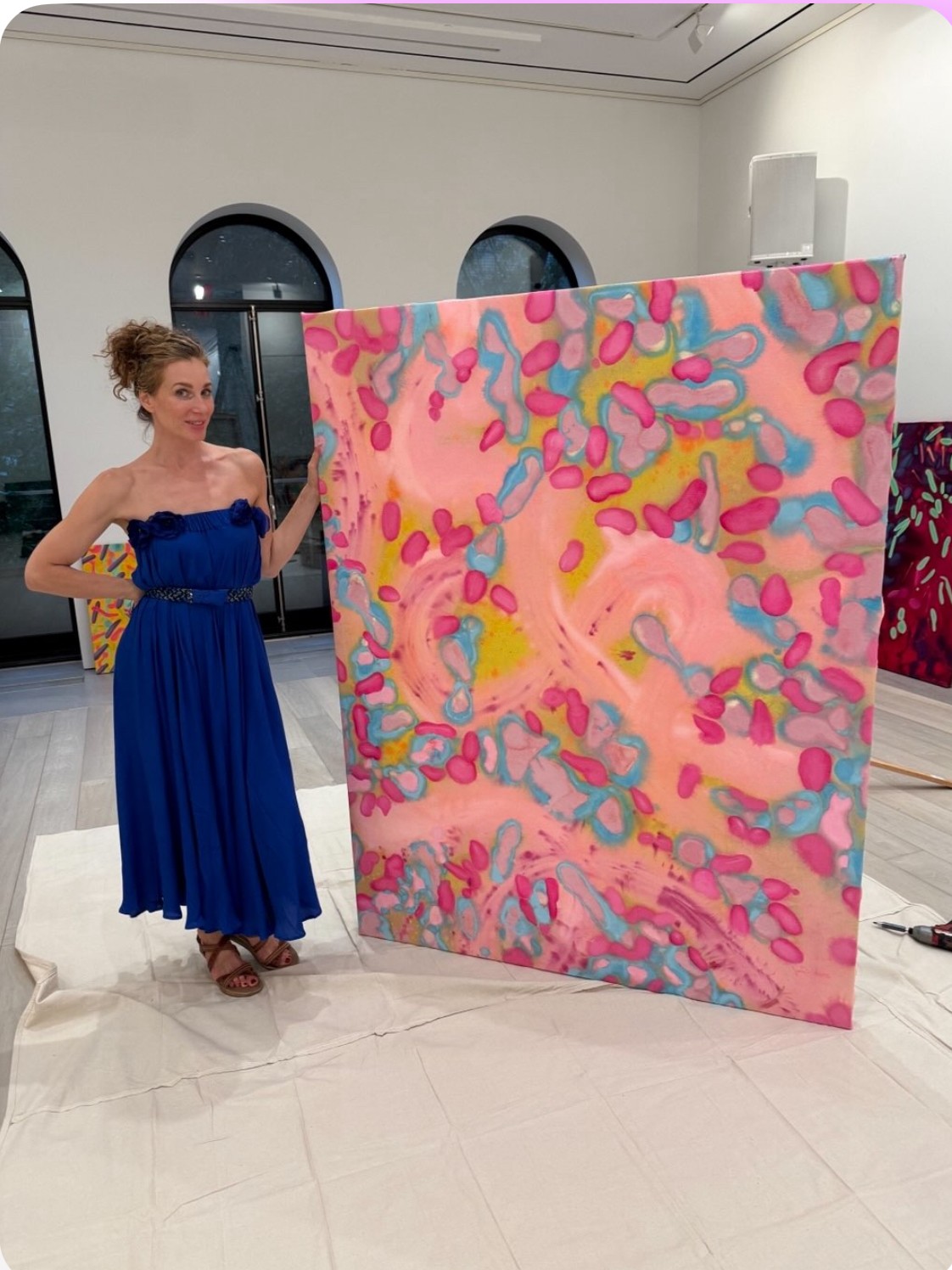 Woman in a blue dress standing next to a colorful abstract painting in an indoor gallery.