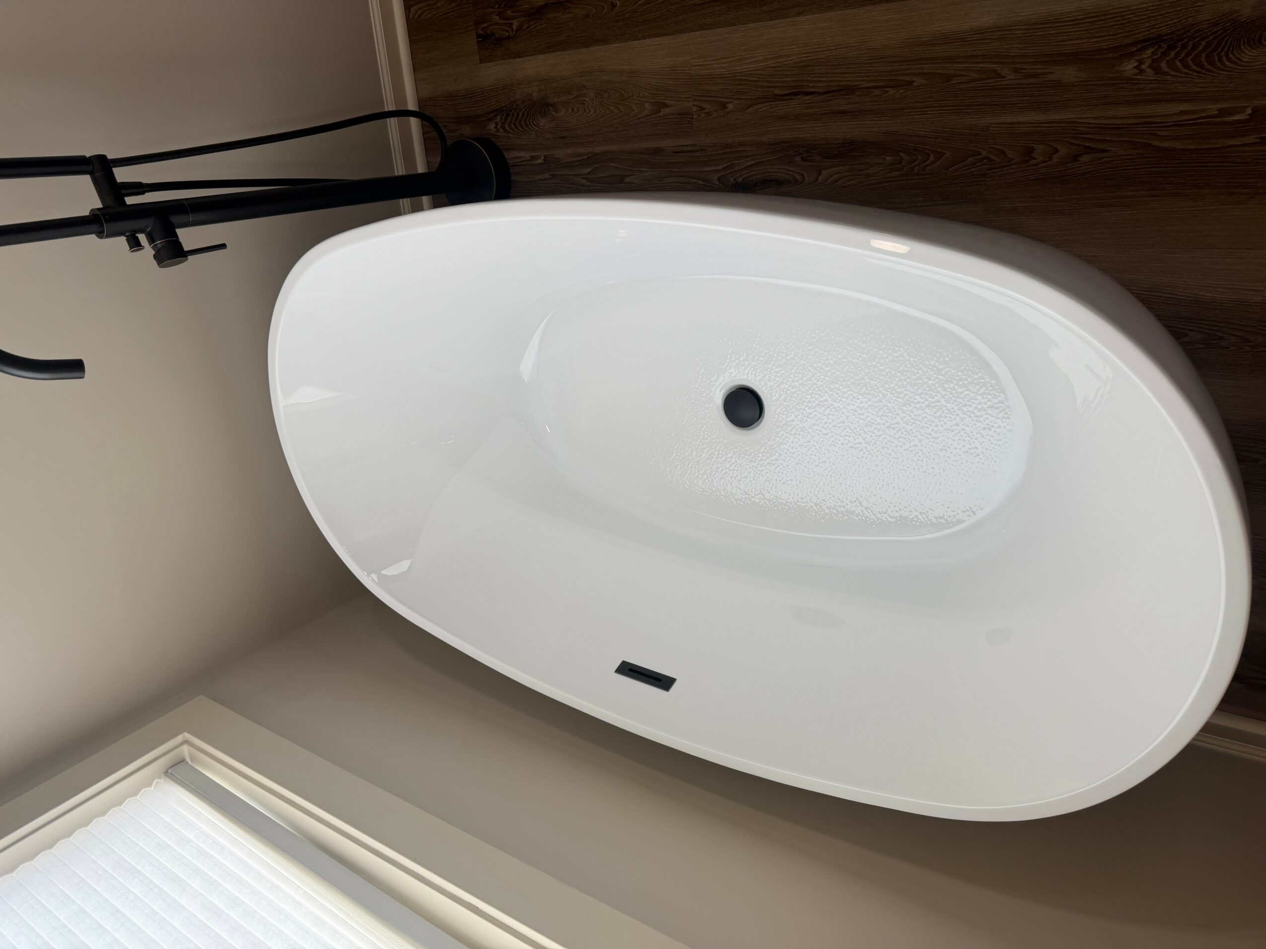 White oval bathtub with drain and faucet, positioned on a wooden floor near a window with blinds.