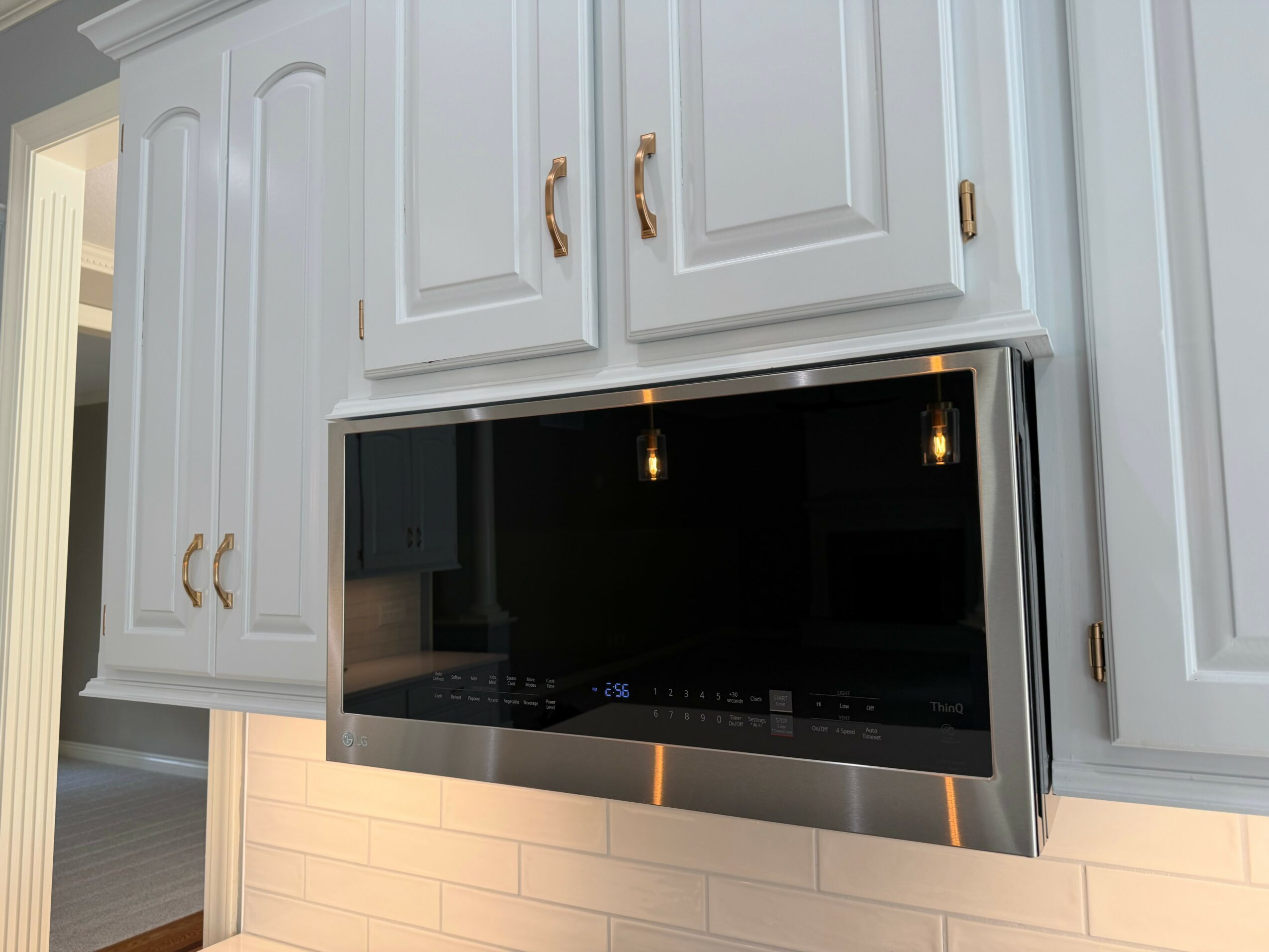 Microwave oven installed above white kitchen cabinets with gold handles, white tiled backsplash, and a partial view of a room.
