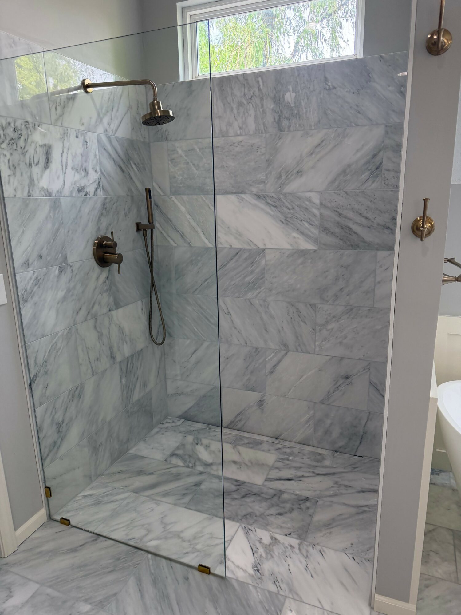 Marble shower with glass door, showerhead, and window, viewed from inside the shower area.