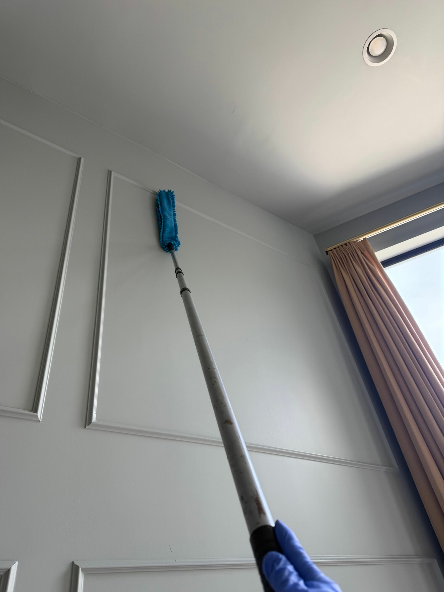 Person cleaning a wall with a long-handled mop near a window, blue glove visible, ceiling light present.