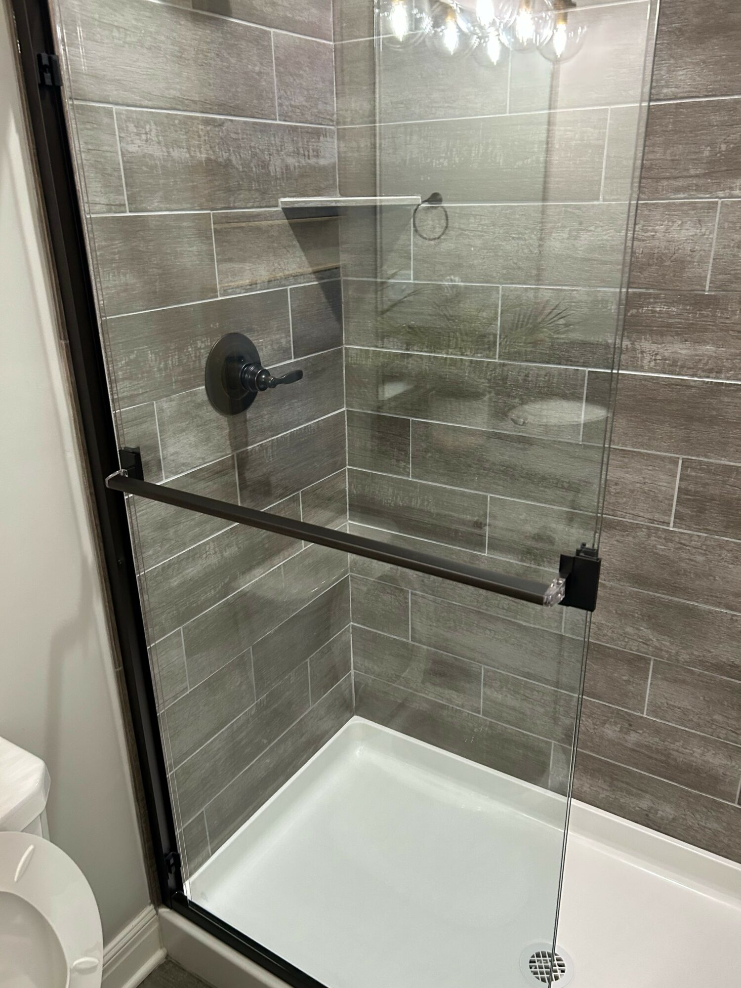 Bathroom shower with glass door, gray tiled walls, and a white bathtub visible in the corner.