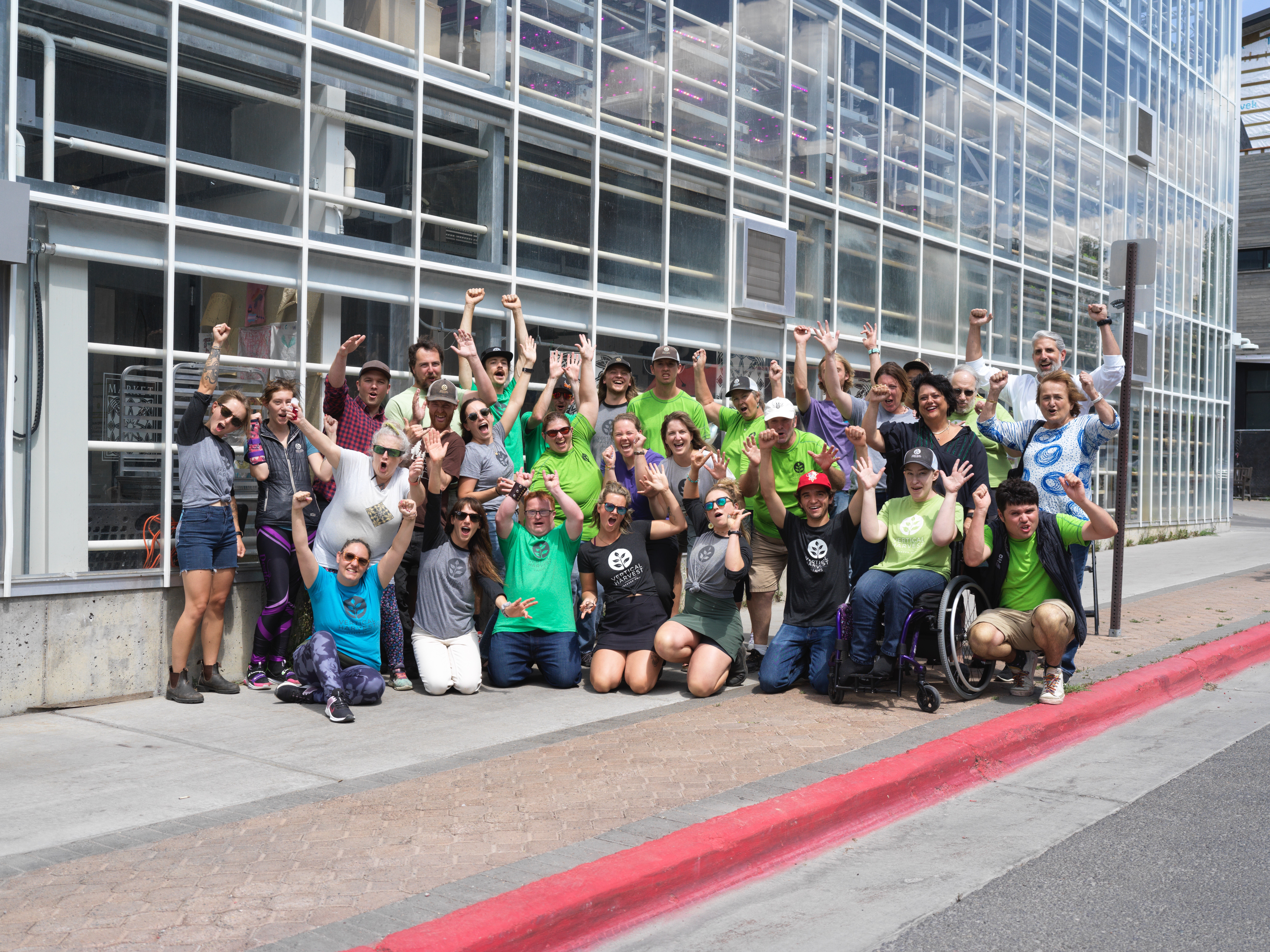Group of people, including children and adults, gathered outside a modern glass building, smiling and raising their hands.