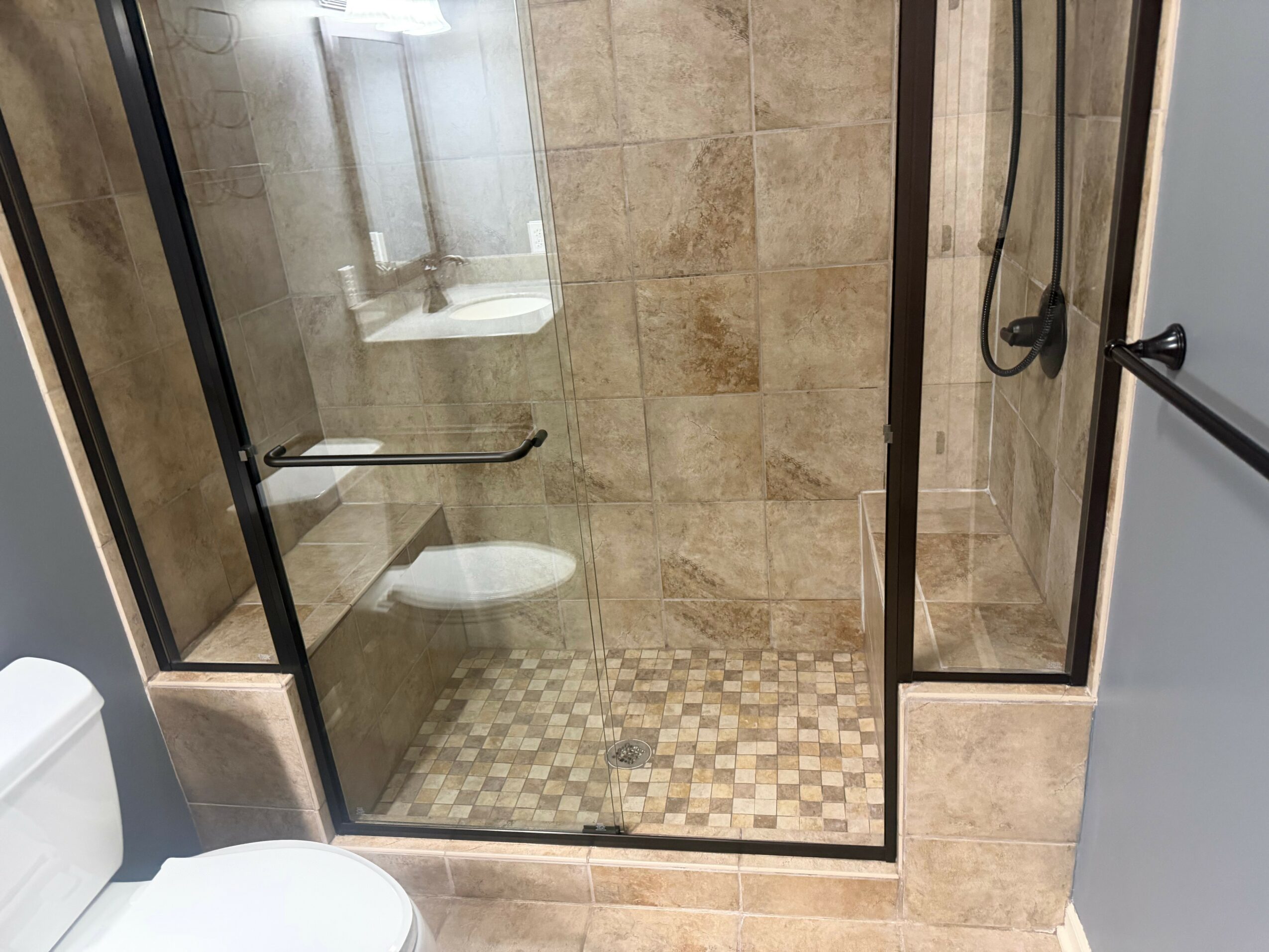 View of a bathroom shower with glass doors, beige tiled walls, and a tiled floor, seen from outside the shower.