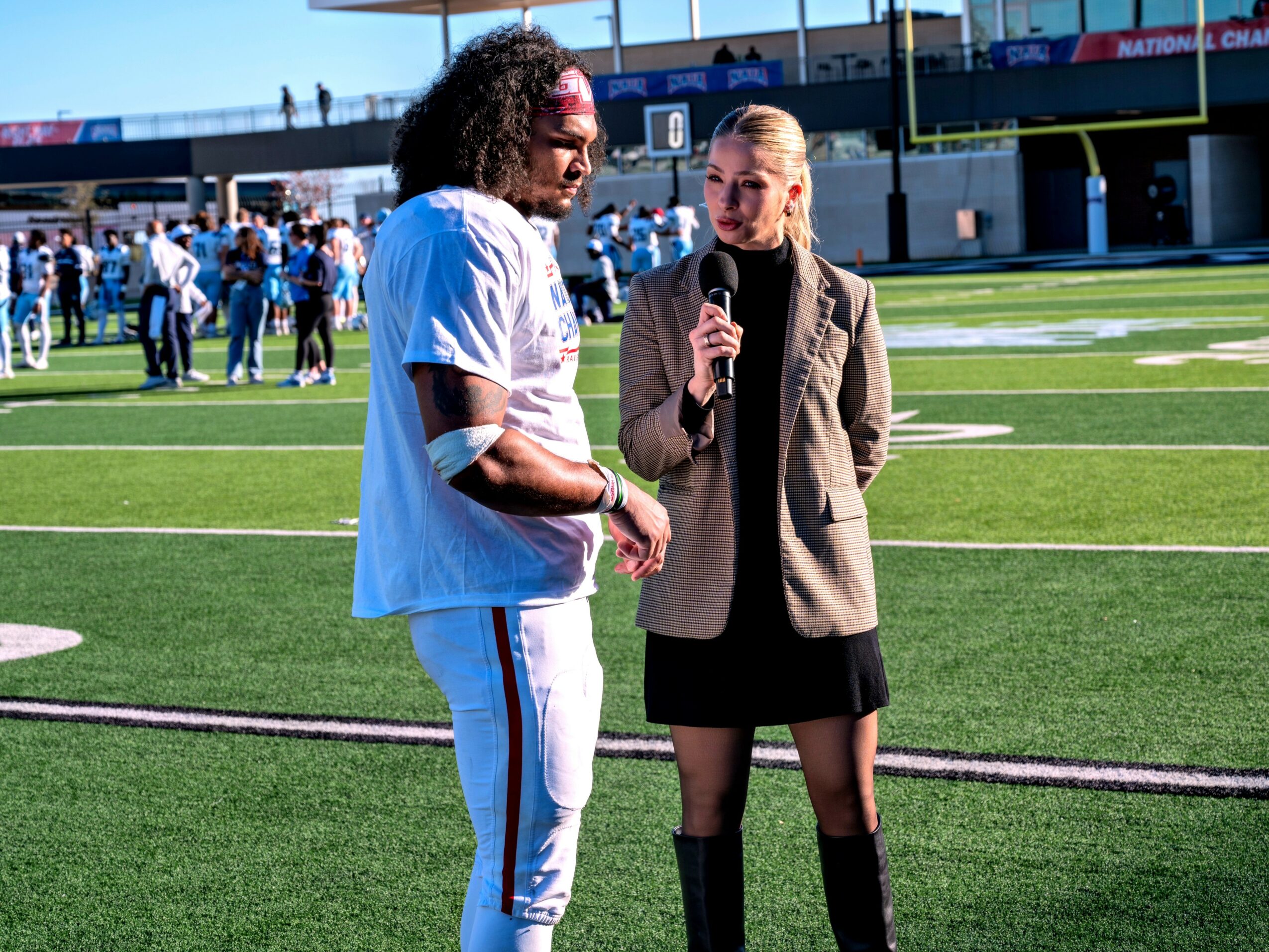 Person in sports uniform talking to a woman with a microphone on a football field, with spectators in the background.