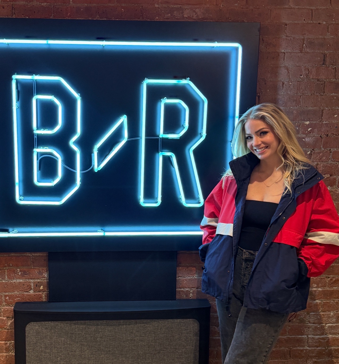 Young woman with blonde hair smiling, wearing a red and navy jacket, standing next to a neon sign with B/R letters, brick wall background.