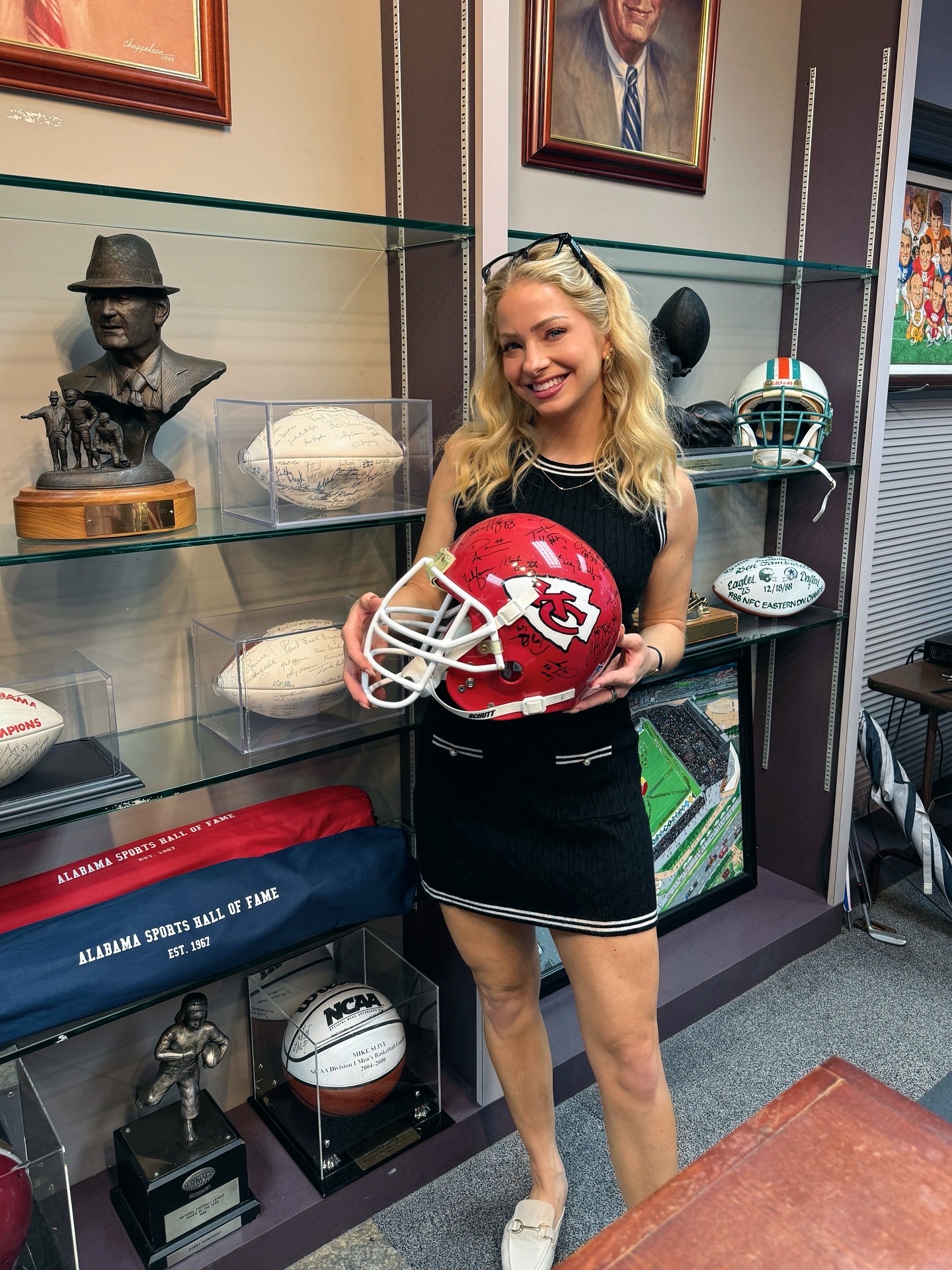Young woman with blonde hair holding a Kansas City Chiefs football helmet, smiling, standing in front of sports memorabilia and framed pictures.