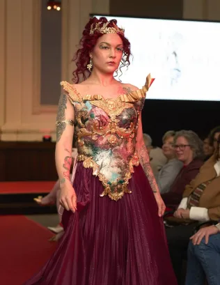 Woman in elaborate dress with floral and gold details, standing in a room with seated audience, facing forward.