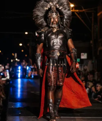 Person in elaborate costume with headdress, armor, and red cape walking on runway at night, audience in background.