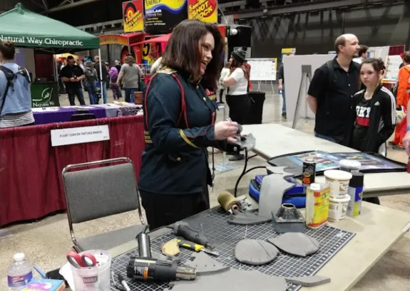 Woman demonstrating tools and equipment at a table in an indoor event space with people in the background.
