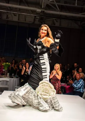 Woman in a black and white dress with a ruffled train, standing on a stage, smiling, with an audience in the background.