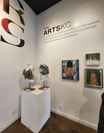 Gallery corner with artwork on white walls, sculptures on pedestal, and a sign reading 'ARTS KC'.