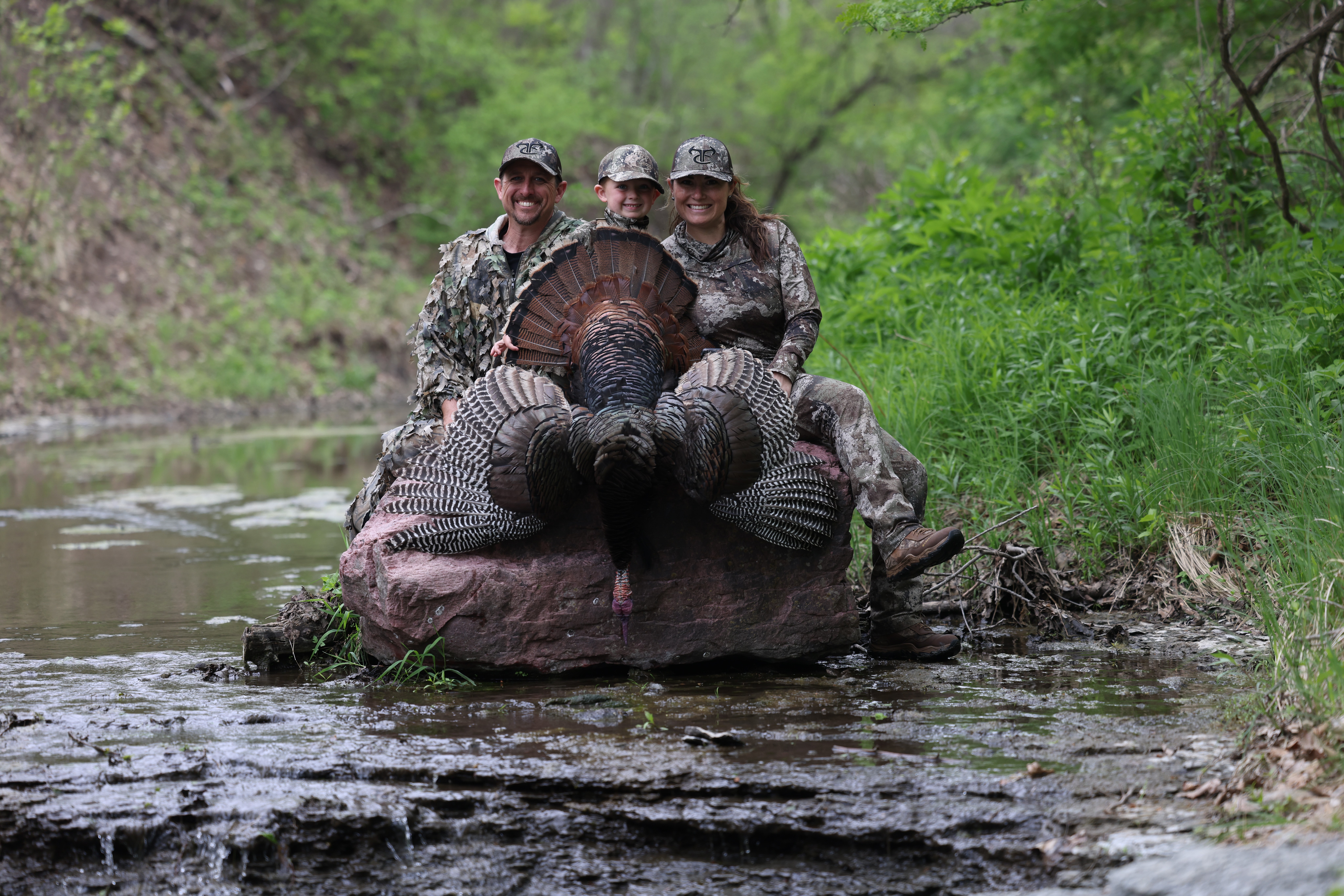 Three people in camouflage clothing sit on a large hippopotamus in a river surrounded by green trees.