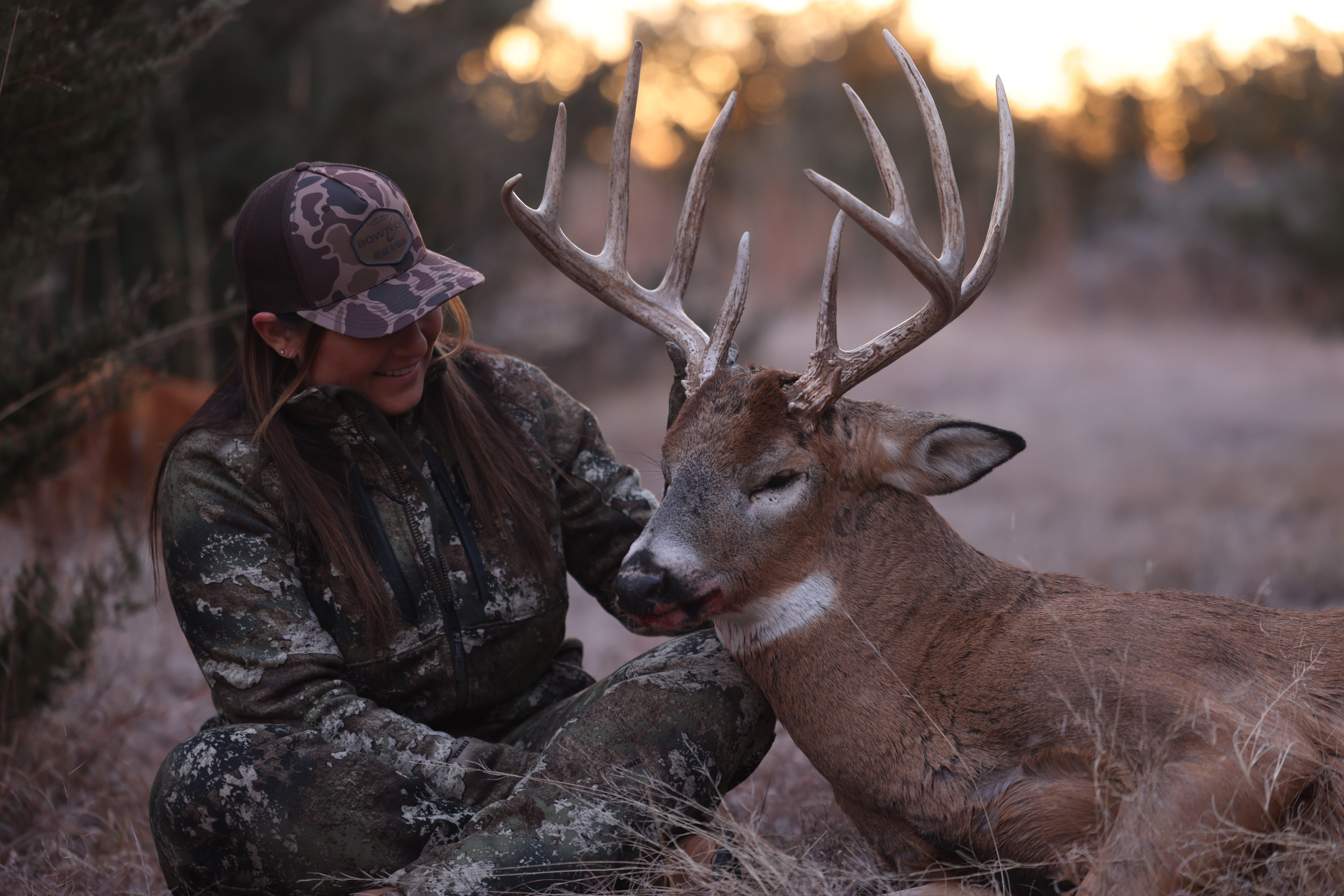 Person in camouflage clothing and hat with a deer in a natural outdoor setting at sunset.