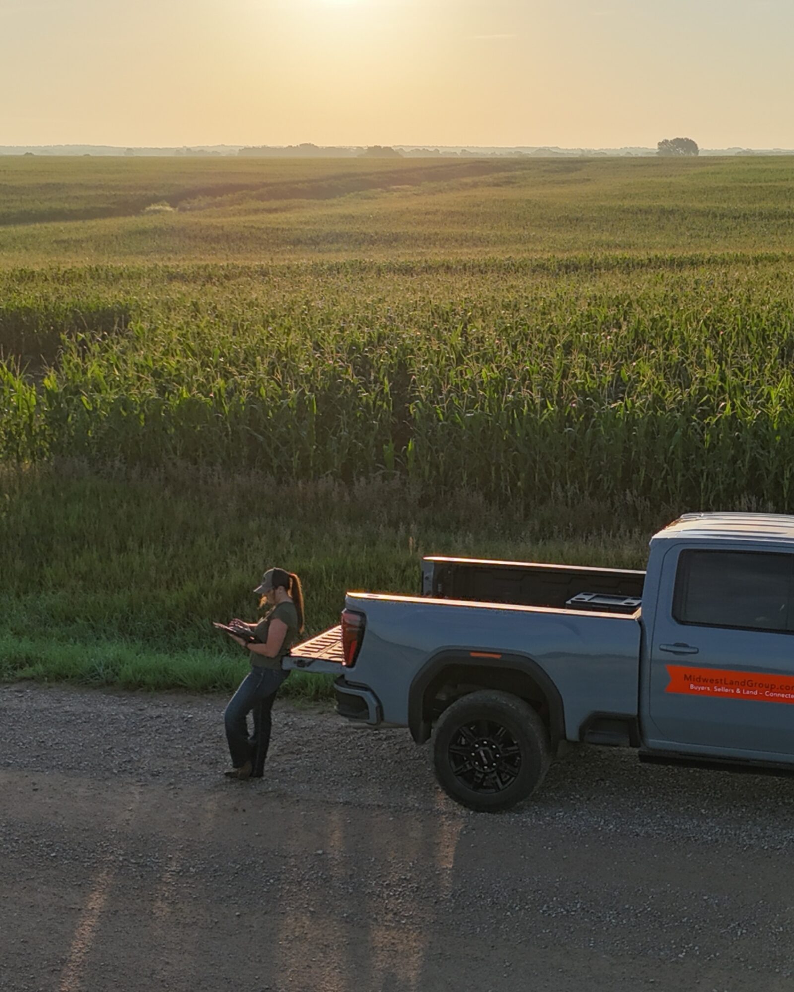 Person standing next to a blue pickup truck on a road, with green fields and a sunset sky in the background.