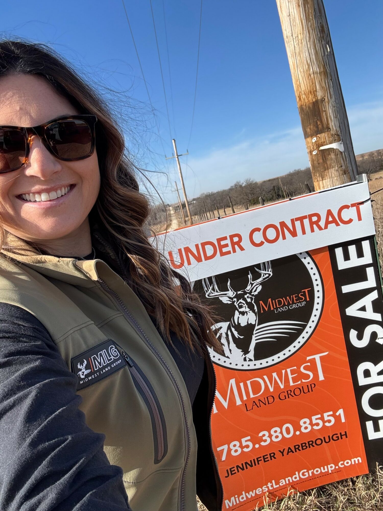 Woman smiling in sunglasses and jacket outdoors, with a sign reading 'UNDER CONTRACT' and 'MIDWEST LAND GROUP' behind her.