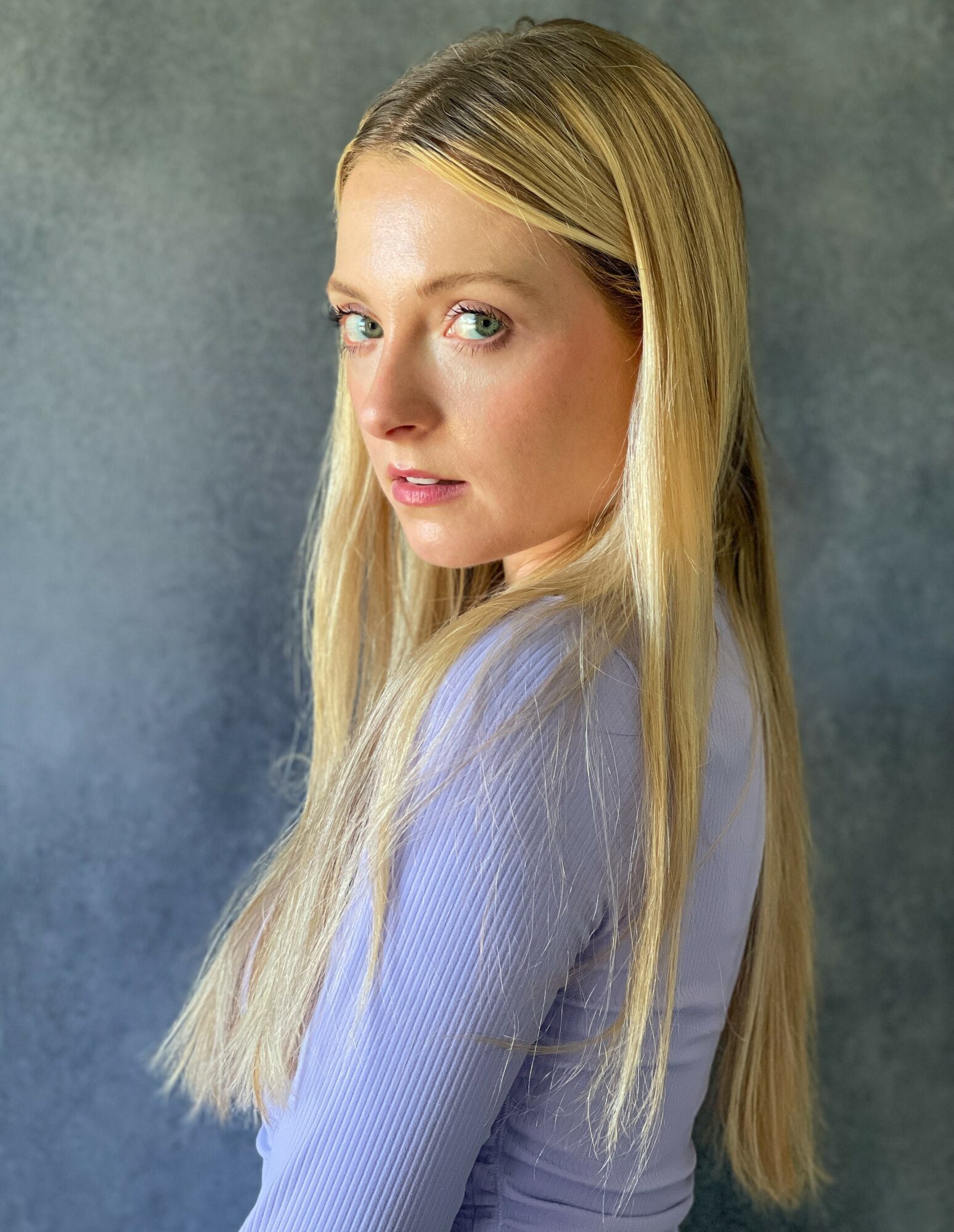 Portrait of a young woman with long blonde hair, looking over her shoulder, wearing a light-colored top, against a textured background.