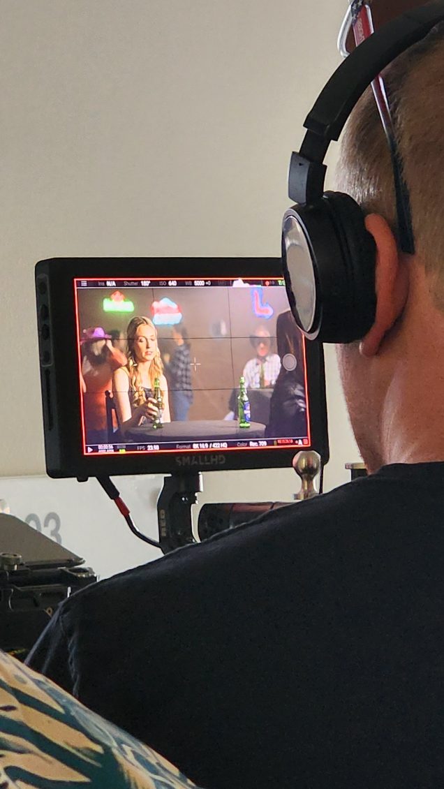 Person wearing headphones looking at a monitor displaying a scene with people, including a woman with curly hair, in an indoor setting.