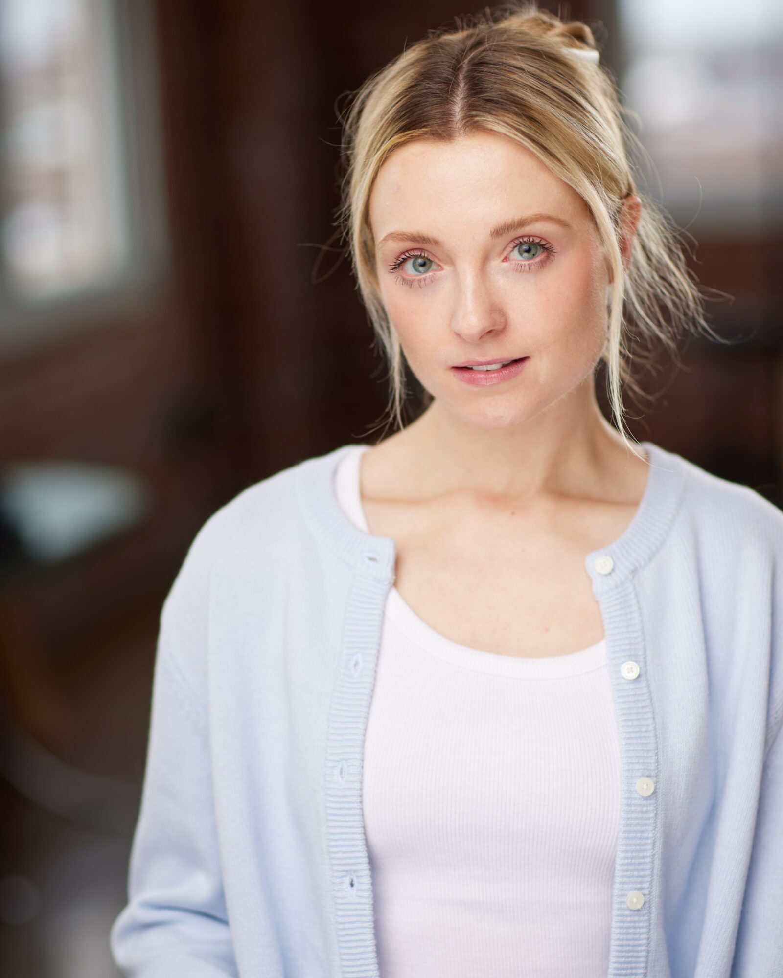 Young woman with blonde hair in a loose bun, wearing a white top and light blue cardigan, indoors with blurred background.