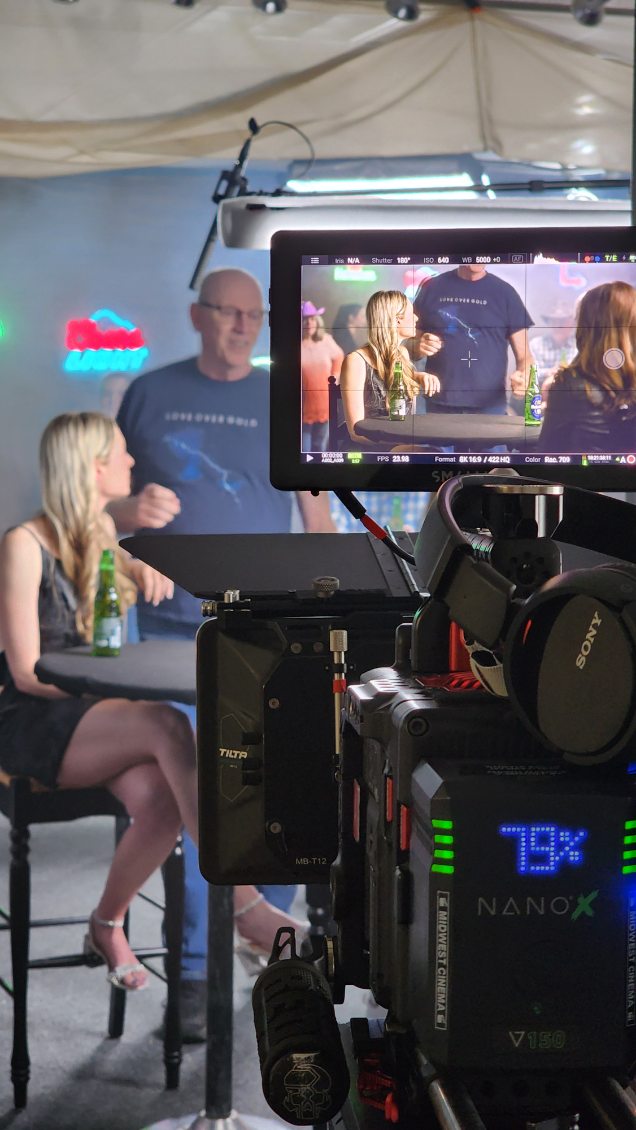 People in a studio setting with a camera and monitor, two women sitting and a man standing, behind a camera setup.