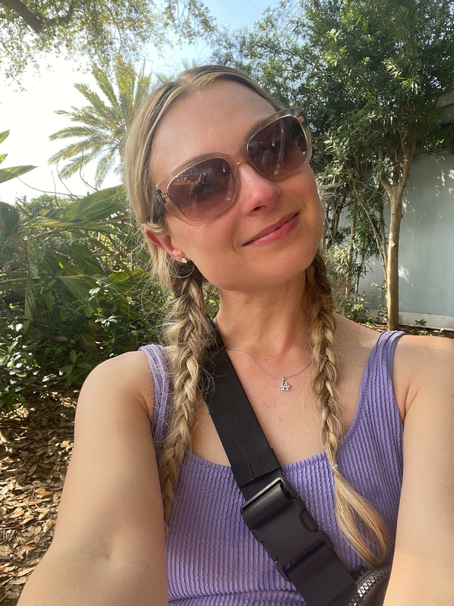 Woman with braided hair wearing sunglasses and a purple top outdoors with trees and plants in background.