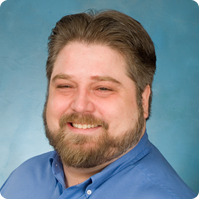Portrait of a smiling man with a beard and short hair, wearing a blue shirt, against a blue background.