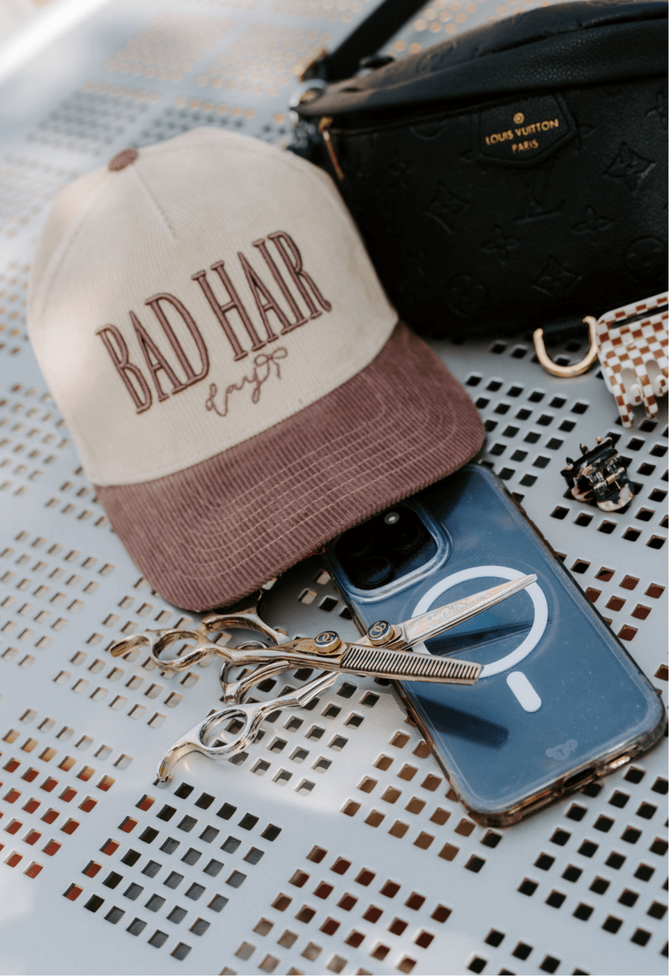 Cap with 'BAD HAIR' text, phone with earphones, black bag, and jewelry on a perforated surface.