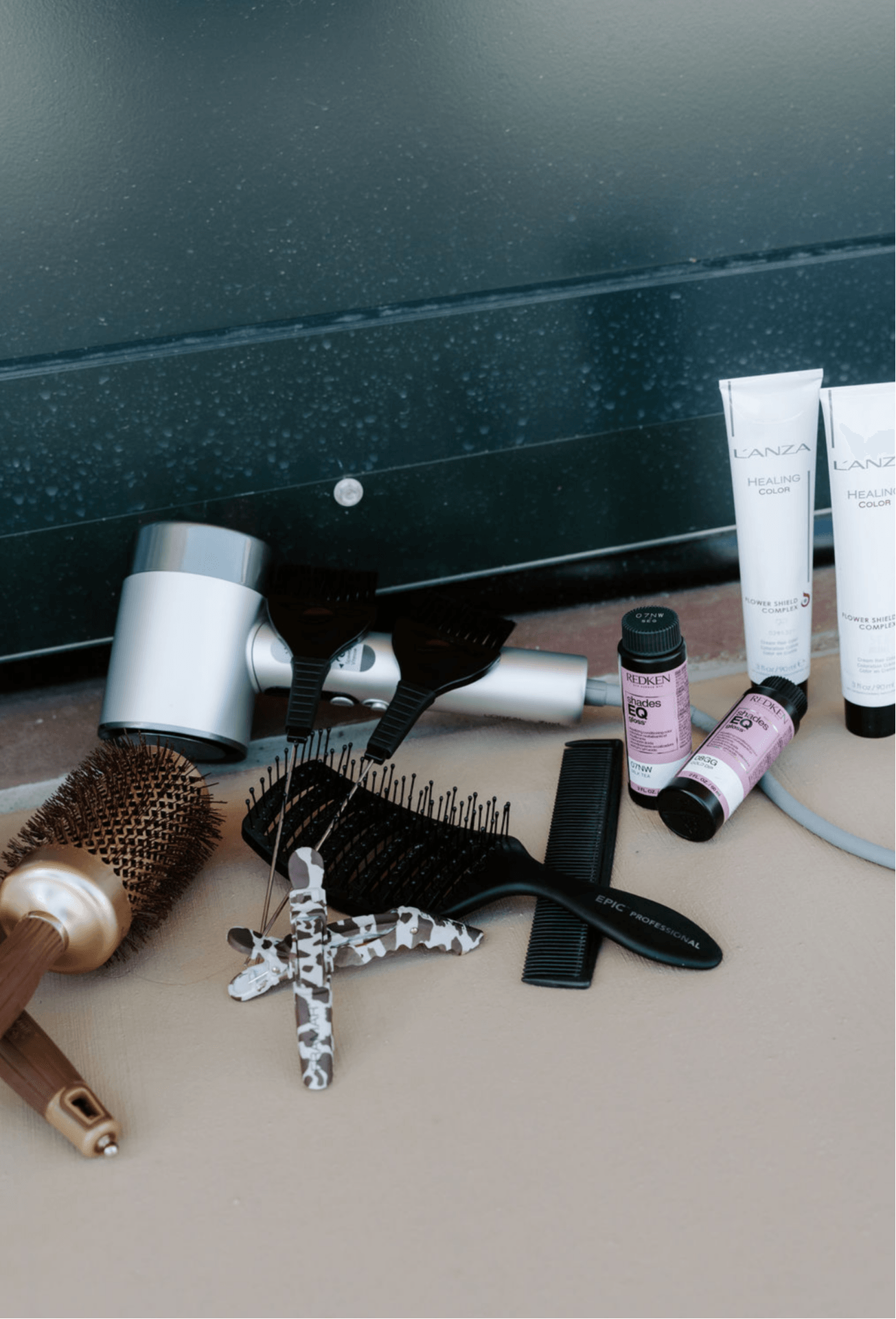 Hairbrush, hairdryer, comb, and hair products on a surface, with a dark background. Items are arranged randomly.