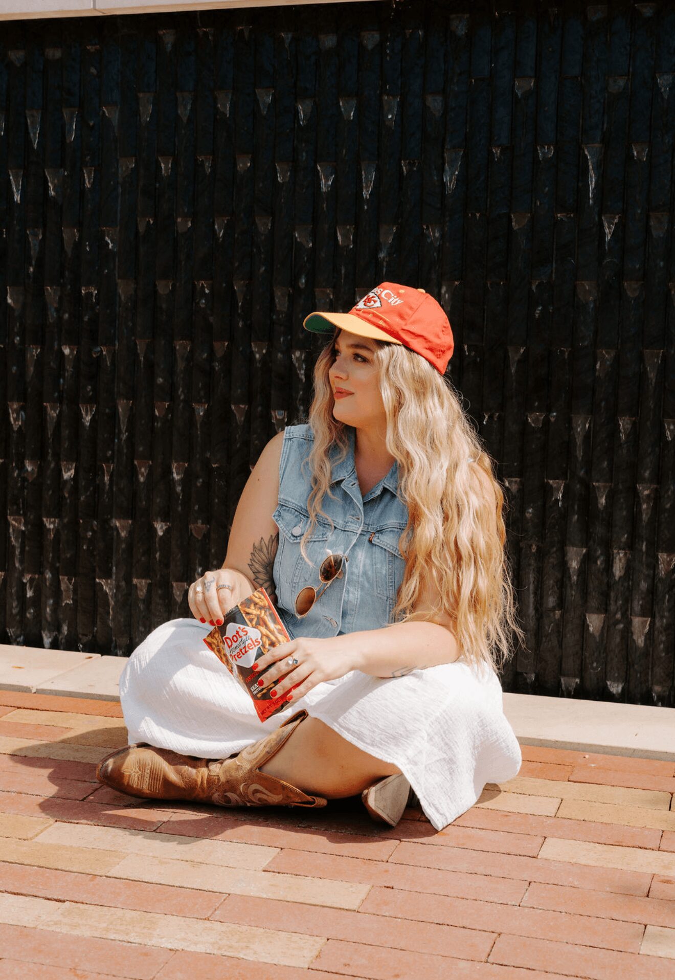 Young woman with long blonde hair sitting on wooden floor holding a drink, wearing a red cap, denim vest, and white skirt.