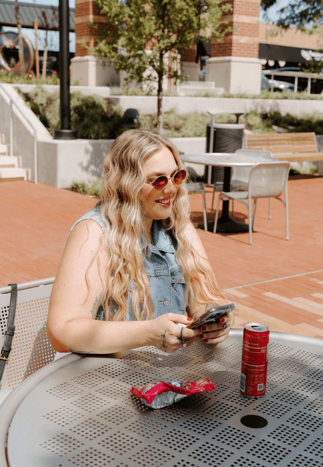 Young woman with long wavy hair and sunglasses sitting at an outdoor table, holding a phone, with snacks and a soda can on the table.