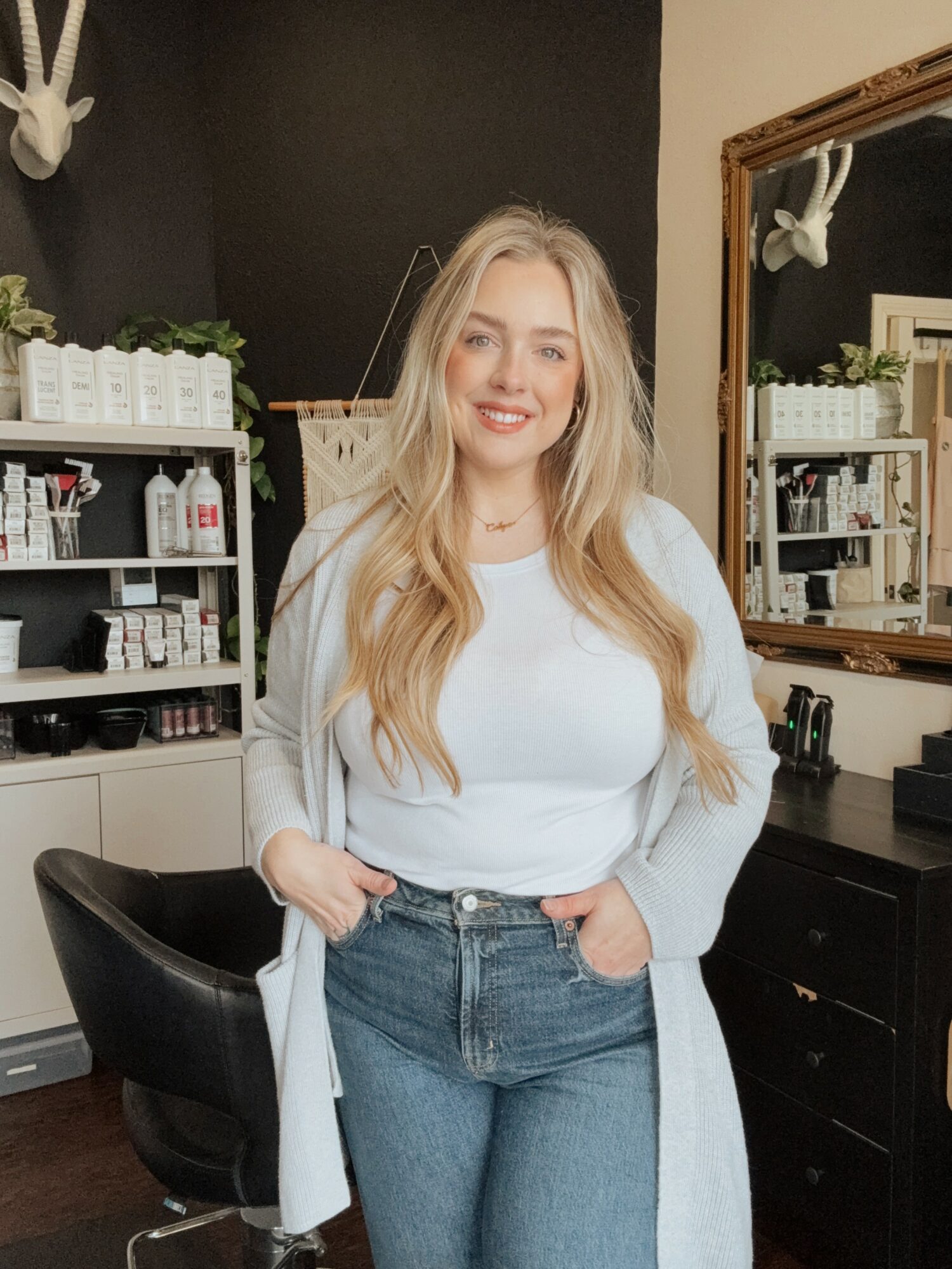 Young woman with long blonde hair smiling, standing in a room with shelves and a mirror, wearing a white top and jeans.