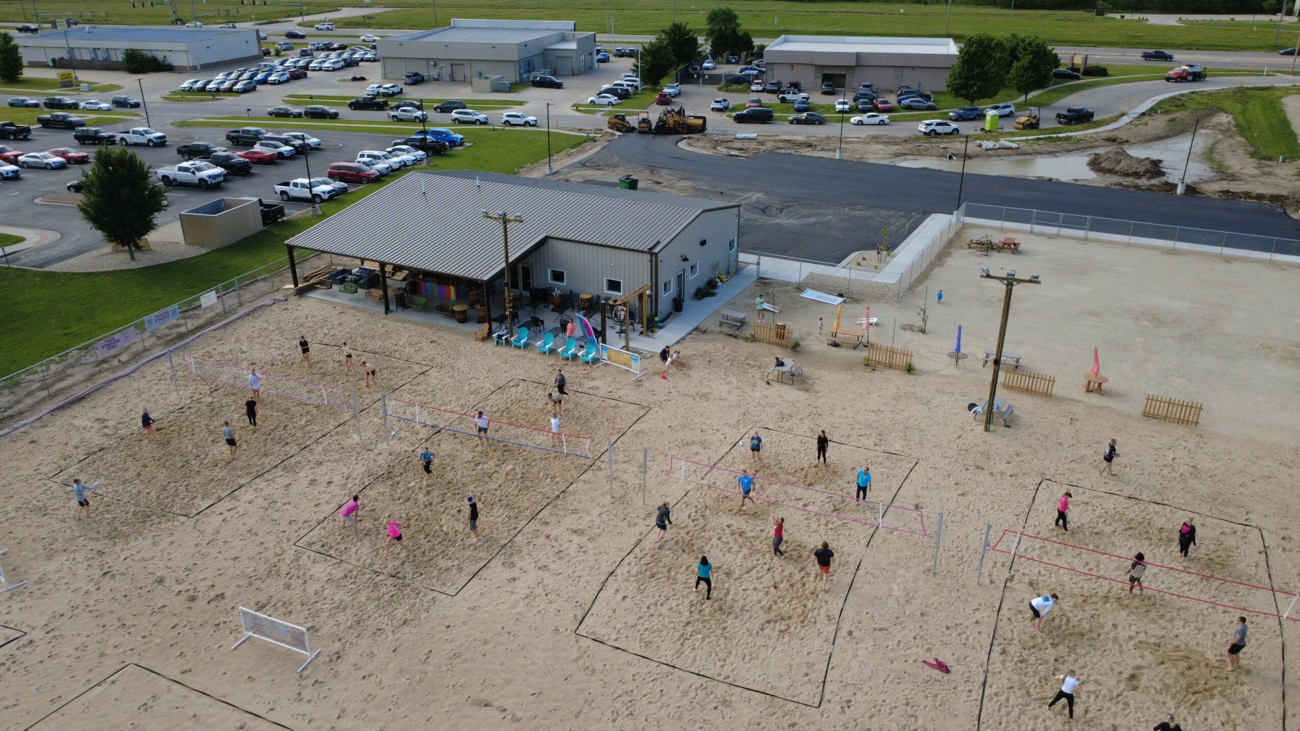 Aerial view of a sandy volleyball court with people playing, adjacent to a building with a parking lot in the background.