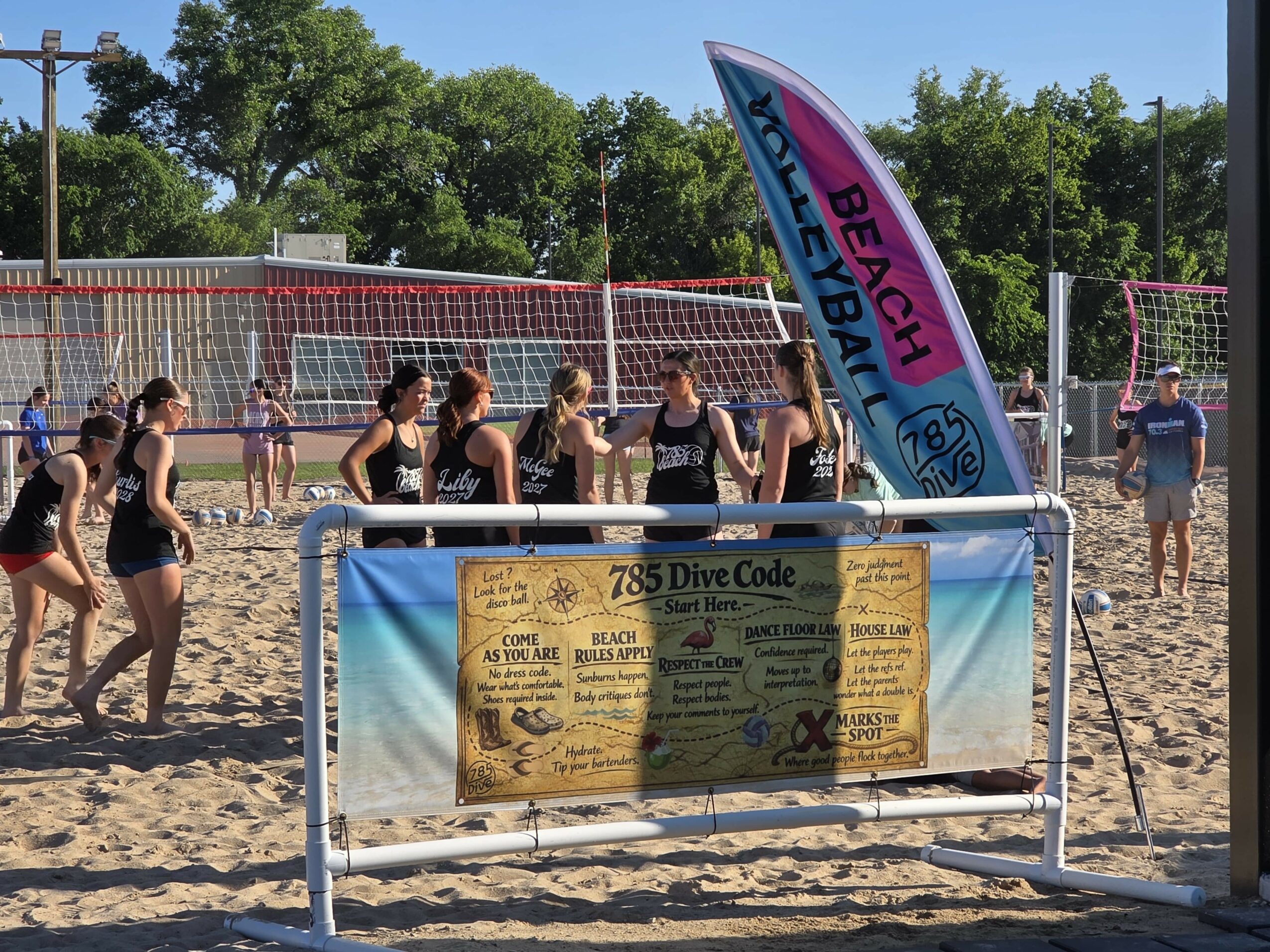 Beach volleyball game with players, a net, and a colorful flag, on sandy court with trees in background.
