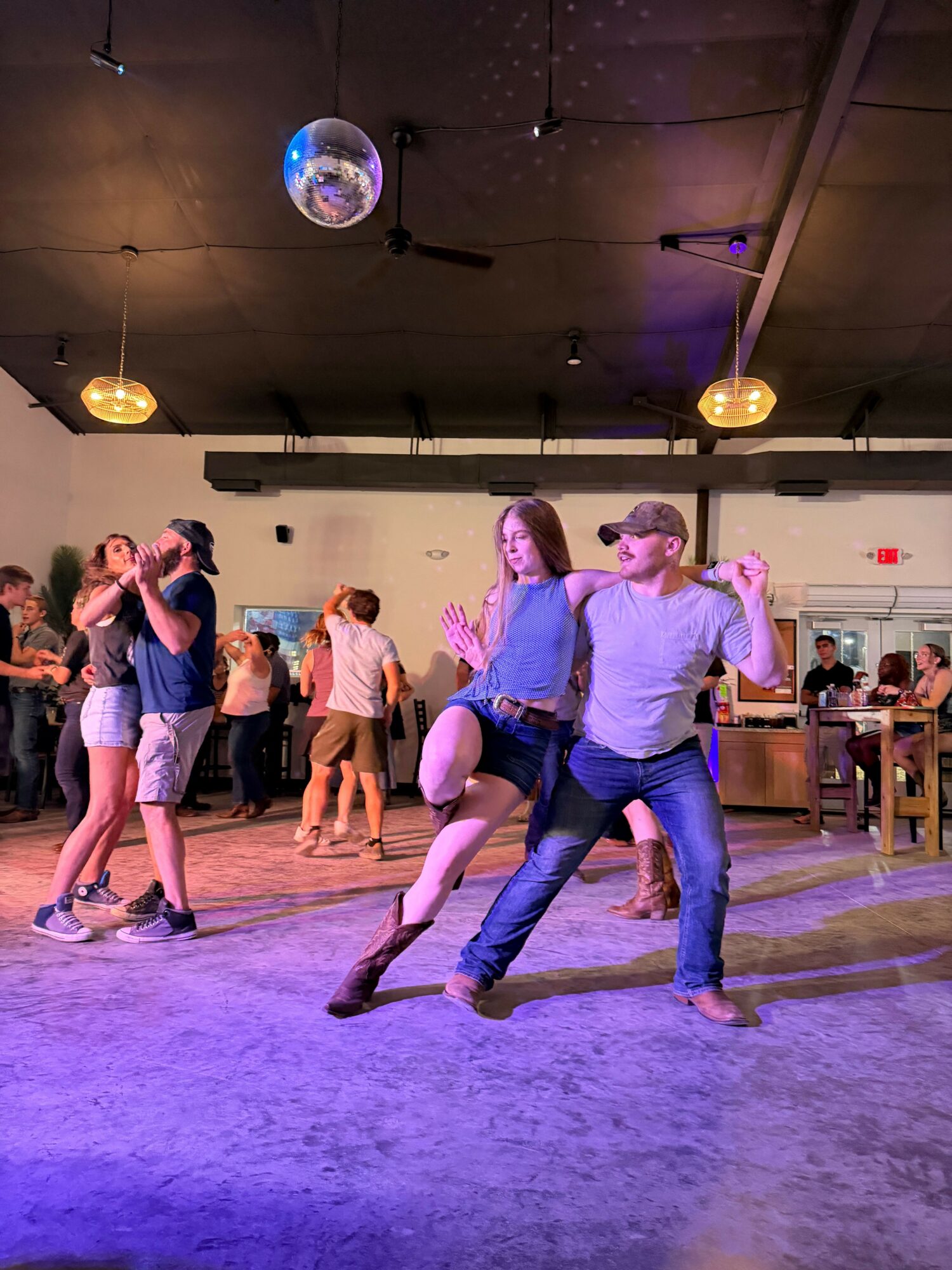 Two women dance together in a dimly lit room with a disco ball, surrounded by other people and wooden furniture.