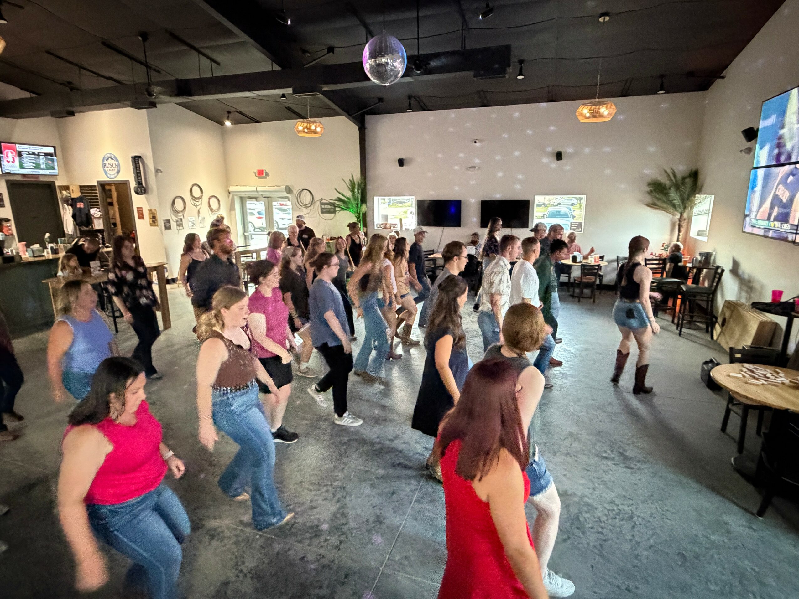 Group of people dancing in a spacious indoor venue with screens and decorations, some watching and others dancing, lively atmosphere.