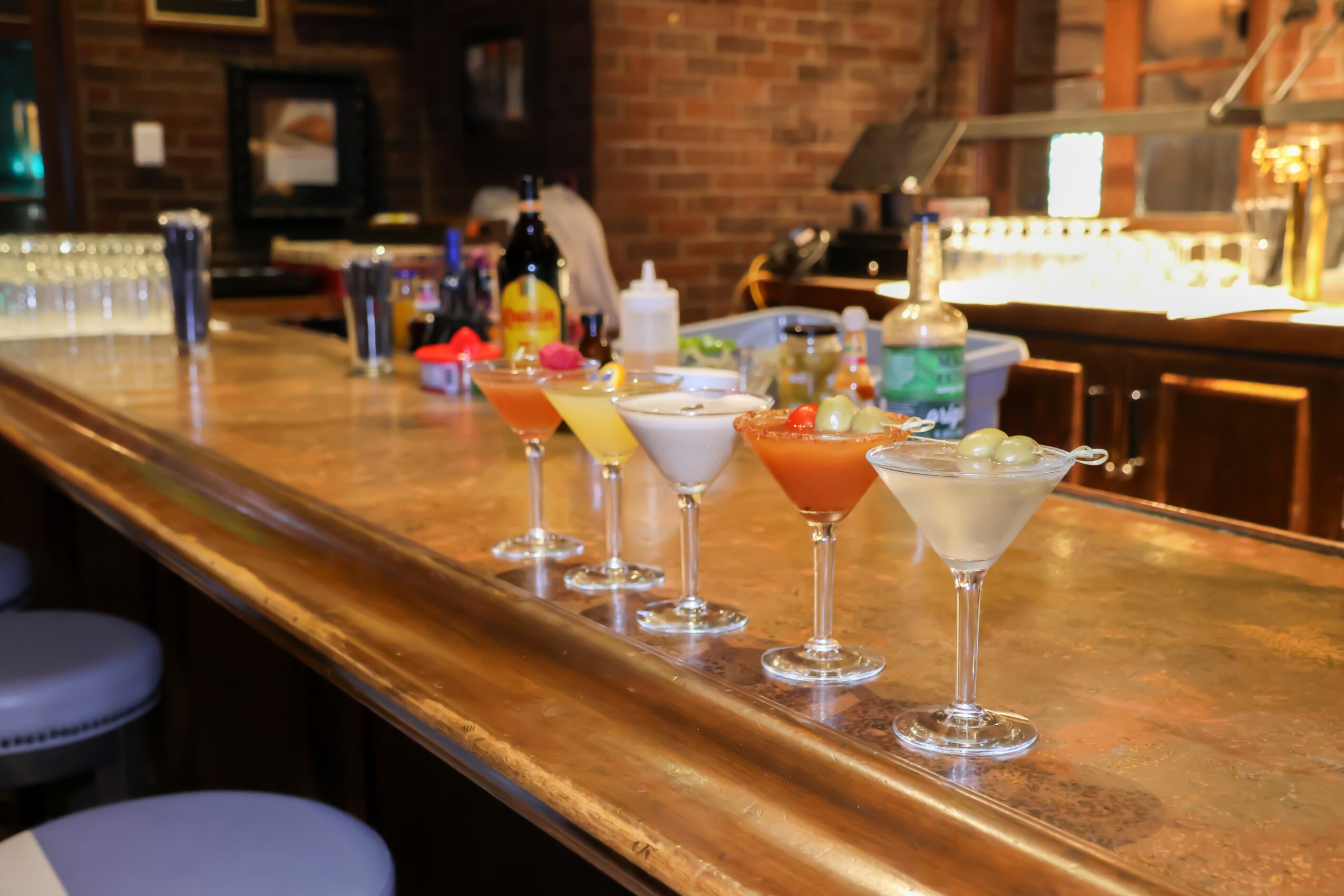 Six colorful cocktails on a bar counter with bottles and bar equipment in background.