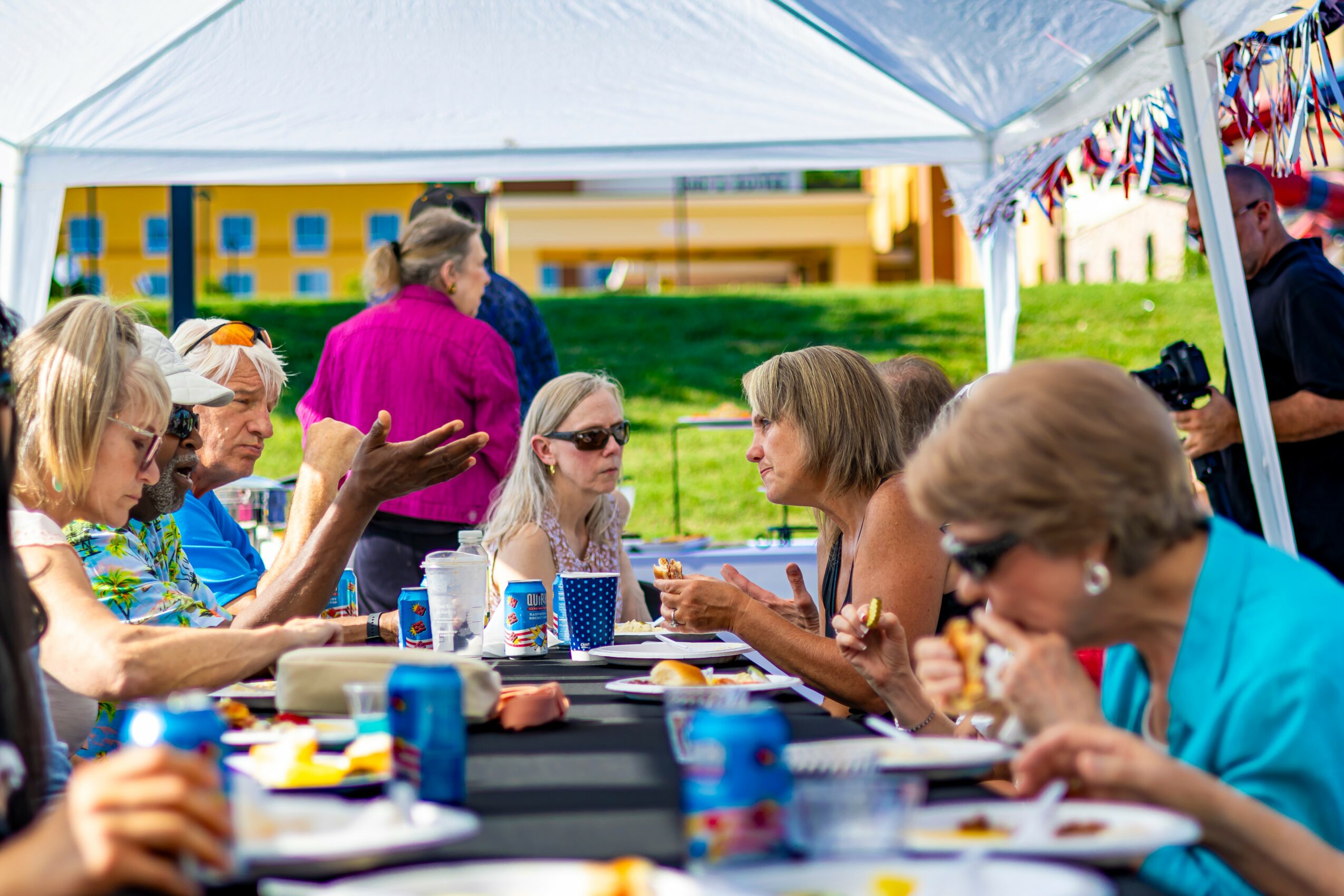 Group of people sitting at a table under a white tent, eating and talking outdoors, with colorful buildings in the background.