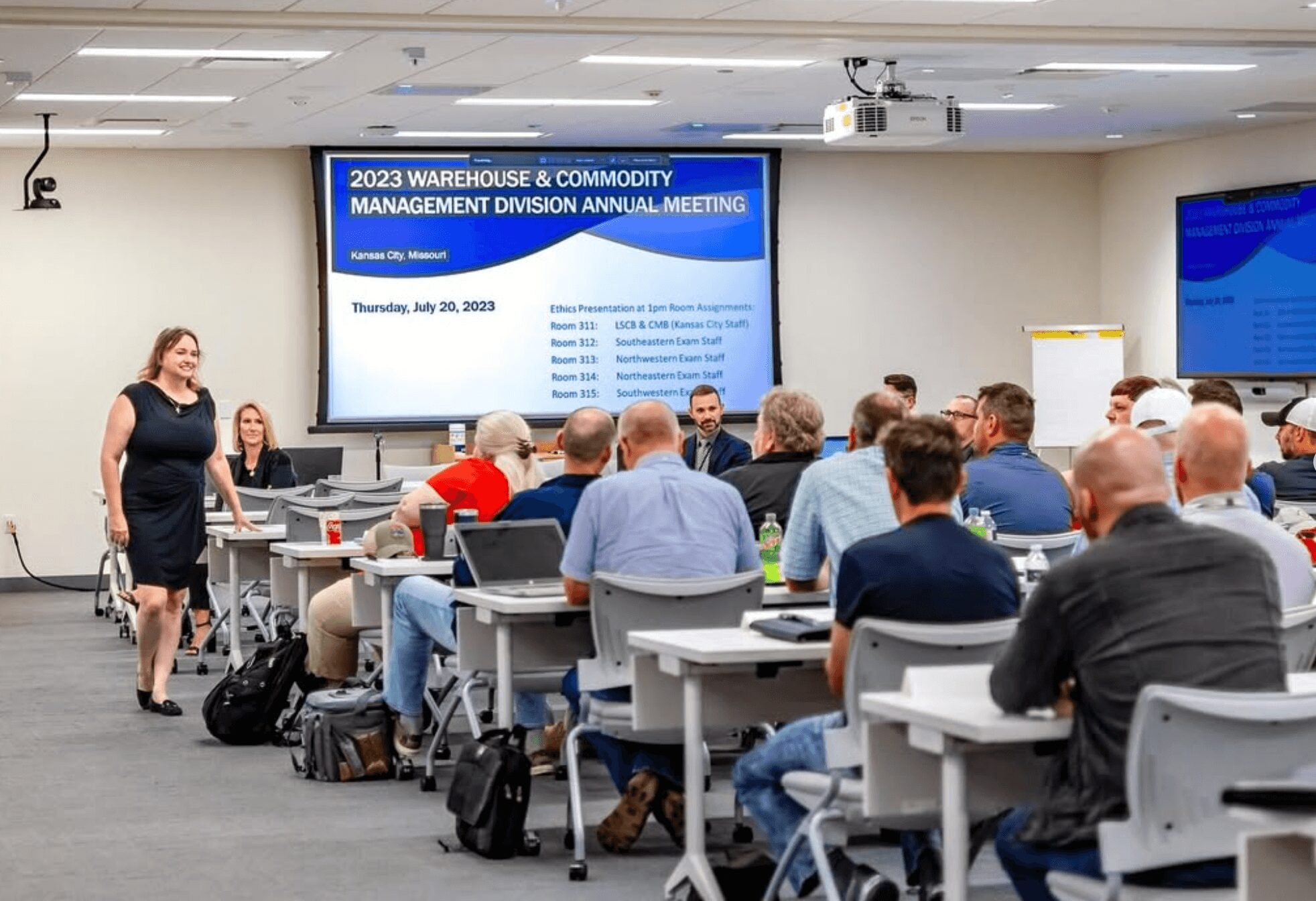 Conference room with attendees listening to a speaker, presentation screens display the event title, date, and agenda.