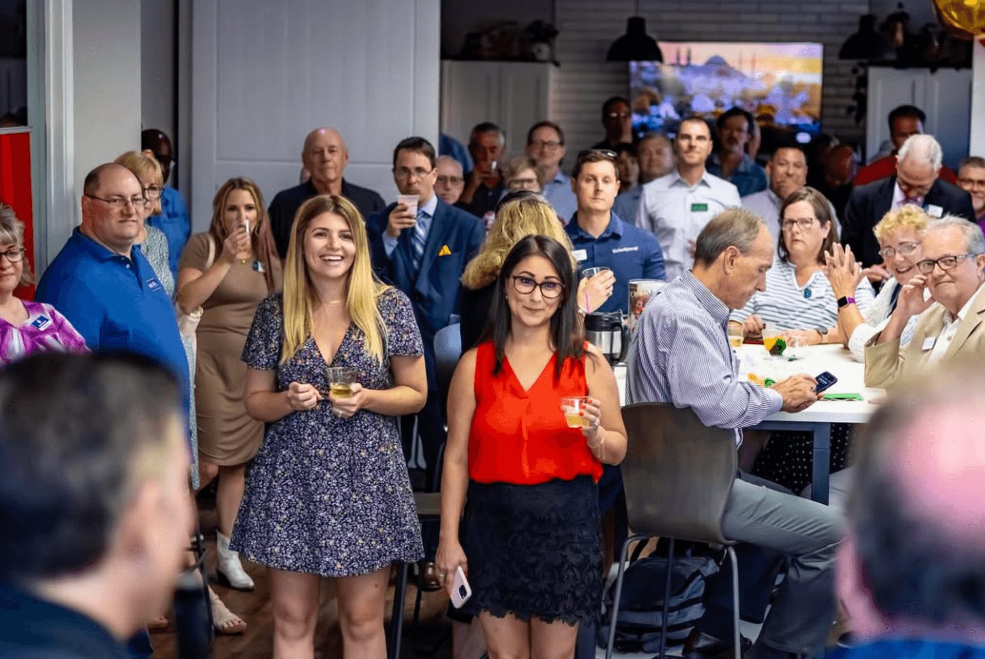 Group of people at a social event, some standing and some seated, with a TV screen in the background.