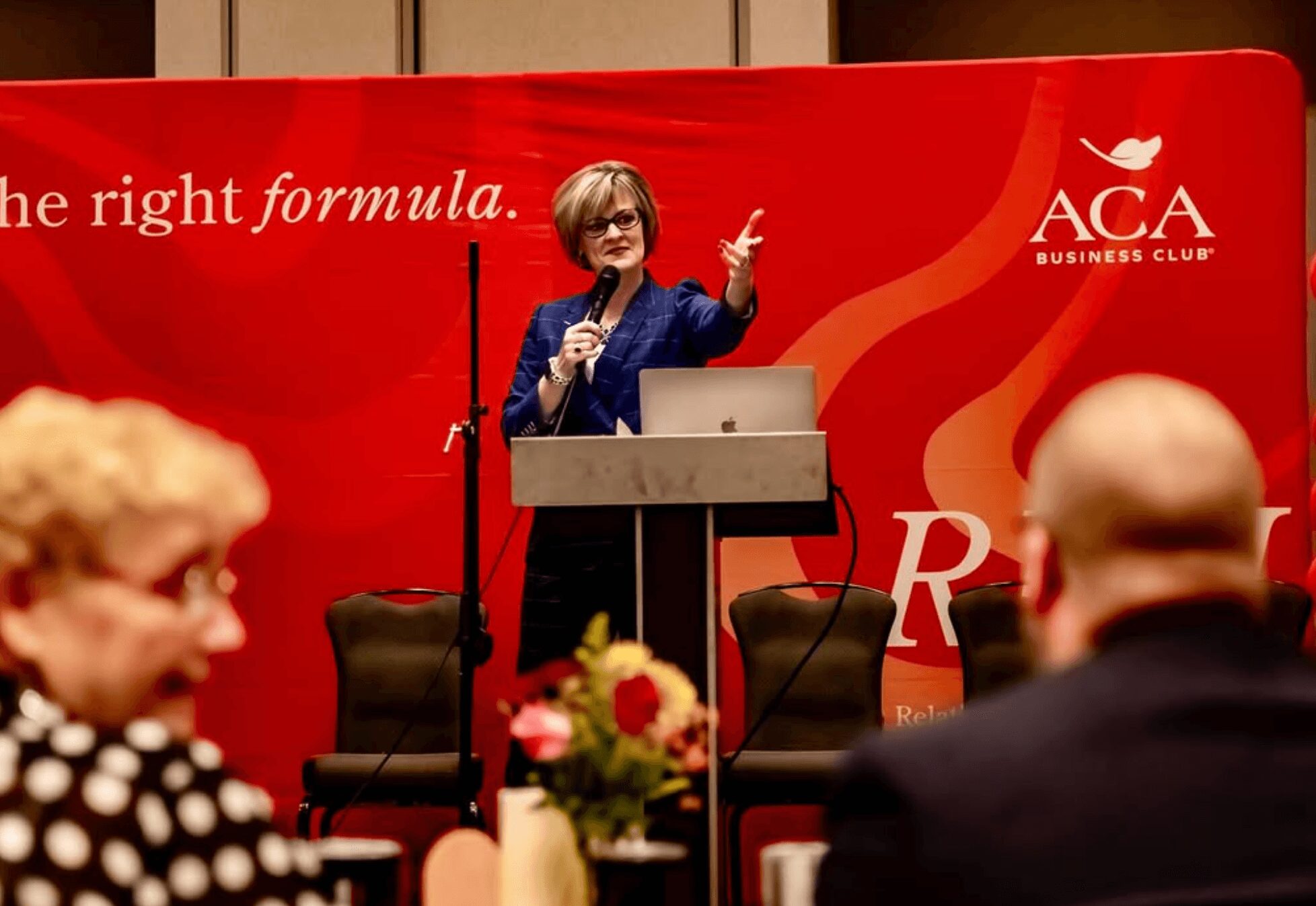 Woman speaking at podium during presentation, with audience in foreground, red backdrop with text and logo, flowers on table.