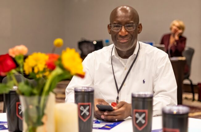 Smiling man in white shirt sitting at table with flowers and drinks, holding a phone, in a social setting.