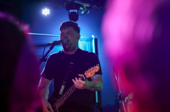 Male musician playing guitar and singing into a microphone on stage with colorful lighting