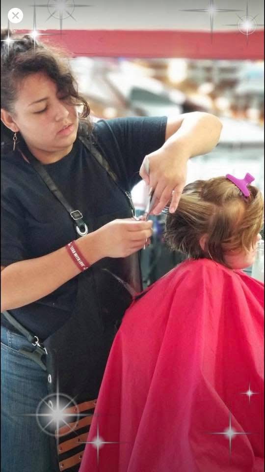 Woman styling a young girl's hair with a pink bow, girl wearing a red cape, in a salon setting.