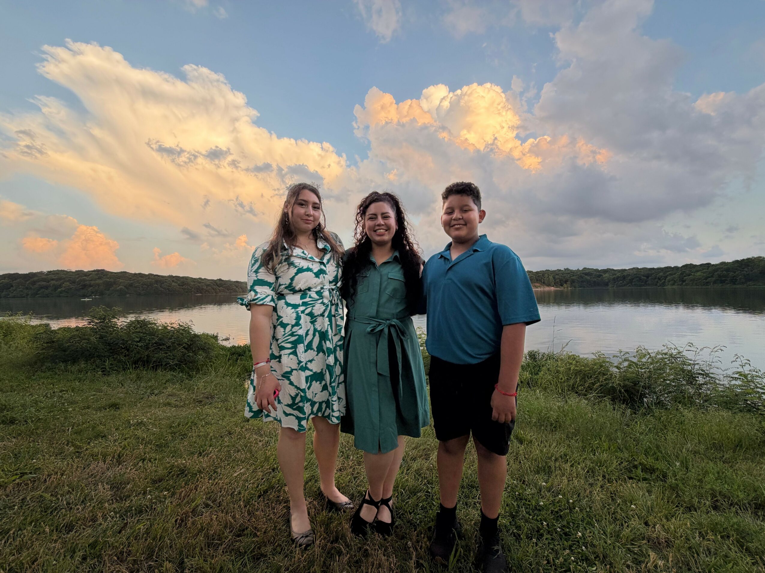 Three people standing outdoors on grass with a cloudy sky background, facing the camera, smiling, with trees in the distance.