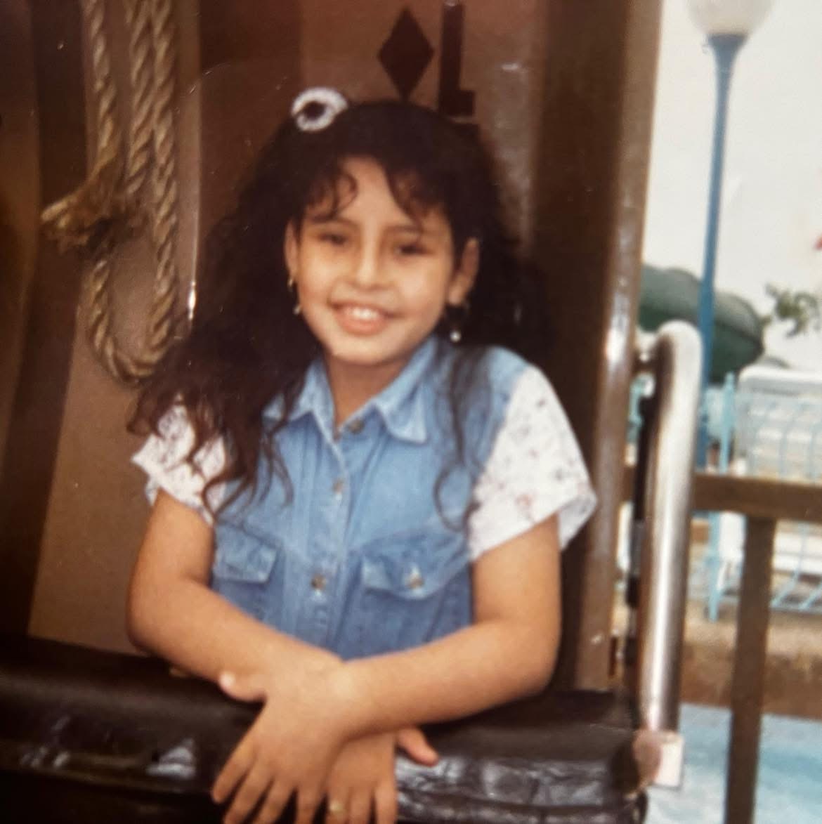 Young girl with curly hair smiling, wearing a denim vest and white shirt, leaning on a black surface.