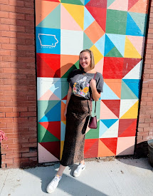 Young woman standing in front of a colorful geometric mural, smiling, wearing a black top, patterned pants, and white sneakers.