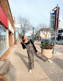 Person standing on sidewalk, leaning back with arms outstretched, wearing patterned jumpsuit and white shoes, in an urban street scene.