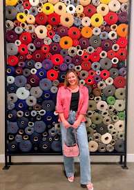 Young woman with pink jacket and jeans standing in front of a colorful wall of yarn spools, smiling.